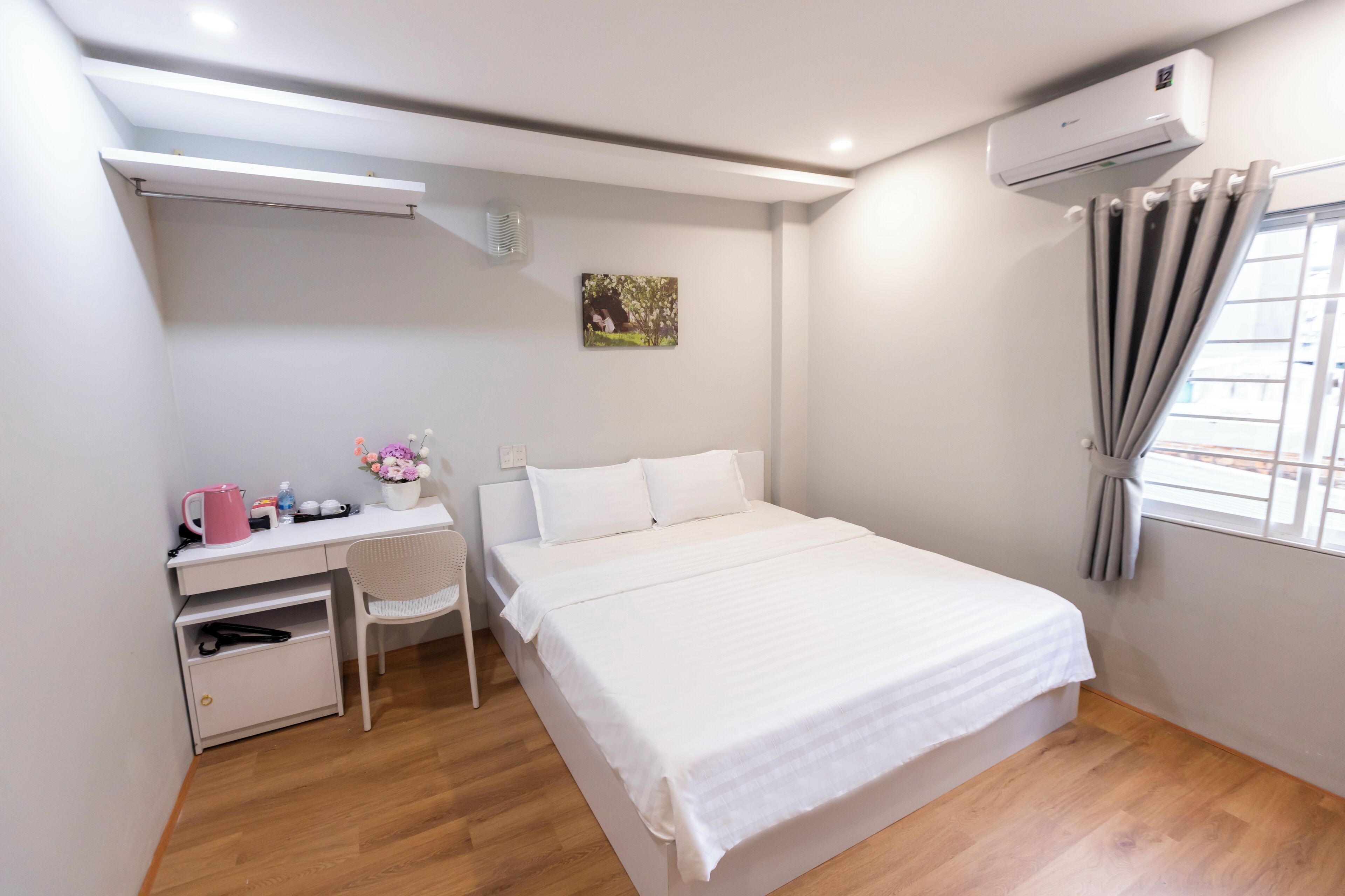 A bedroom with a bed, a desk, a chair, a small table with a pink kettle, a picture on the wall, and an air conditioner.