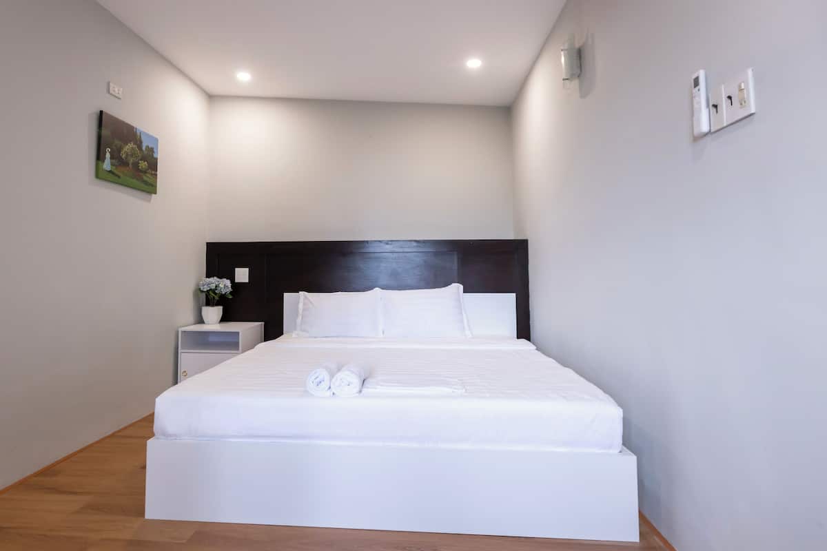 A neatly made bed with white linens and rolled towels, a bedside table with a vase of flowers, and a painting on the wall.