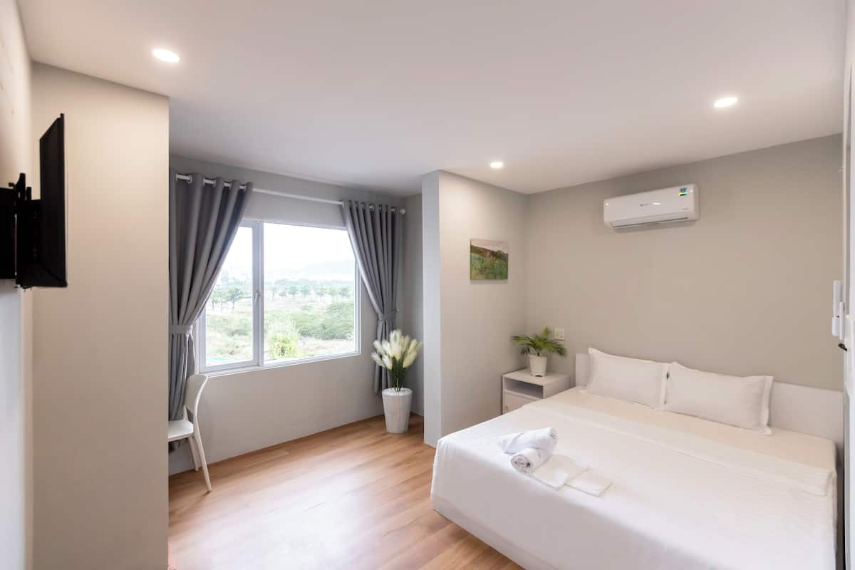 A modern bedroom with a large bed, a bedside table with a plant, a window with curtains, and a mounted air conditioner.