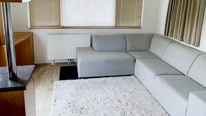 Una sala de estar moderna con un sofá seccional gris, un tapete blanco con textura, un radiador y una ventana con persianas.