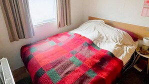 Una habitación con cama, ventana con cortinas, mesita de noche con lámpara y estante con libros.