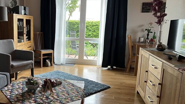 A living room with a wooden cabinet, a television, and a view of the outdoors through a large window. - Holiday Apartment Alleblas (Geldern)