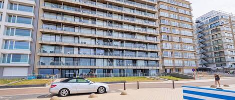 Un immeuble résidentiel à plusieurs étages doté de balcons, d’une zone pavée avec un banc et d’une voiture garée.