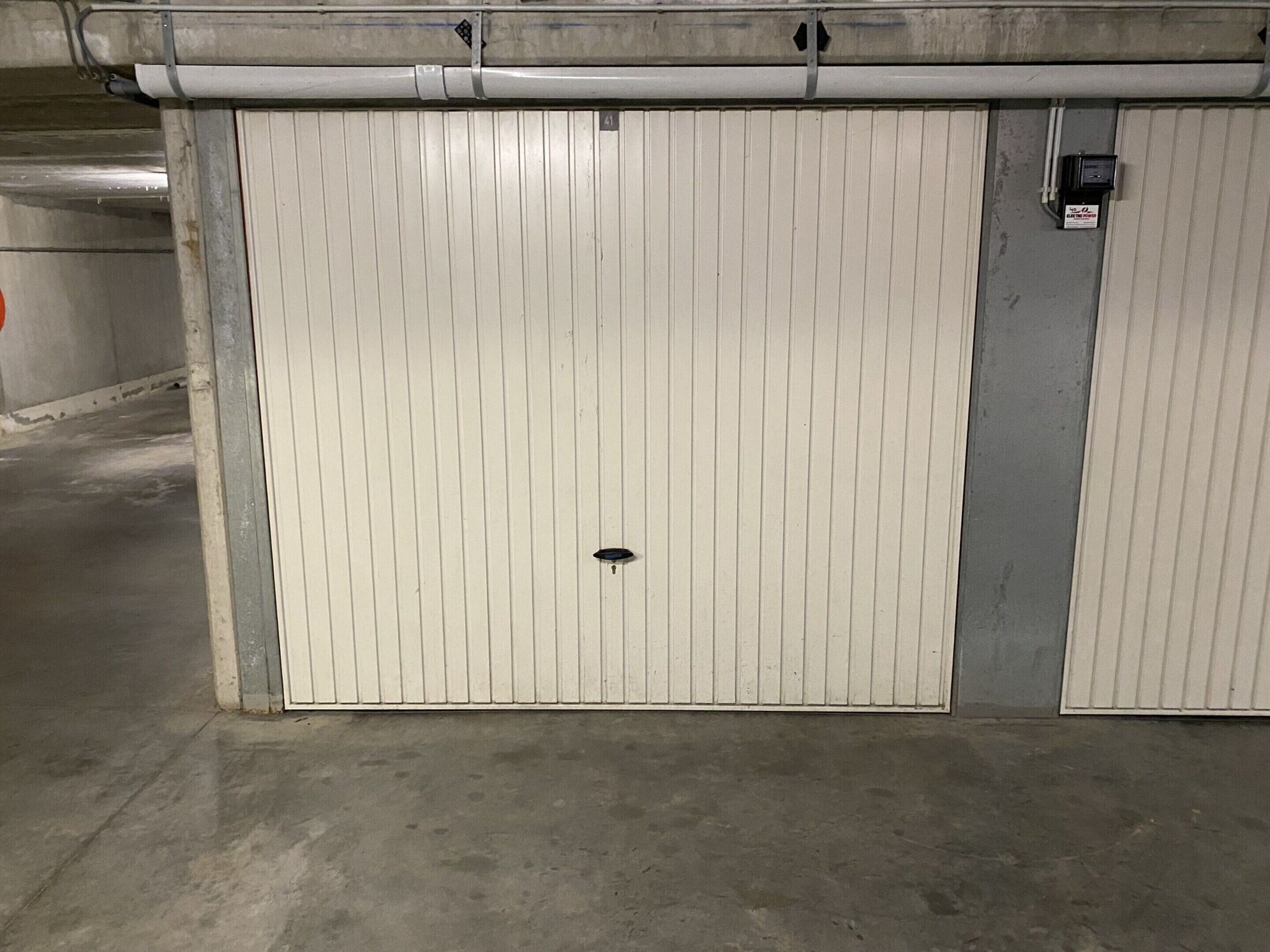A closed, white, double garage door with a small black lock in the center, set in a concrete floor with a metal structure above.