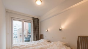 A bedroom with two beds, a window with curtains, a wooden bench, and wall-mounted lights. - Apartment in Nieuwpoort by the Seafront (Koksijde)