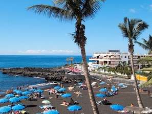 A beach with numerous blue umbrellas, palm trees, and a building with a sign that reads "Hotel". - Belvilla by OYO Apartment in Guaa de Isora (Guia de Isora)