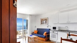 A compact living space with a kitchenette, a dining area, and a sofa with a view of the sea.