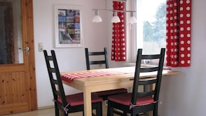 Een eetruimte met een houten tafel en vier zwarte stoelen met rode kussens. Daarboven hangen twee hanglampen en er is een raam met een rood-wit stippen gordijn.