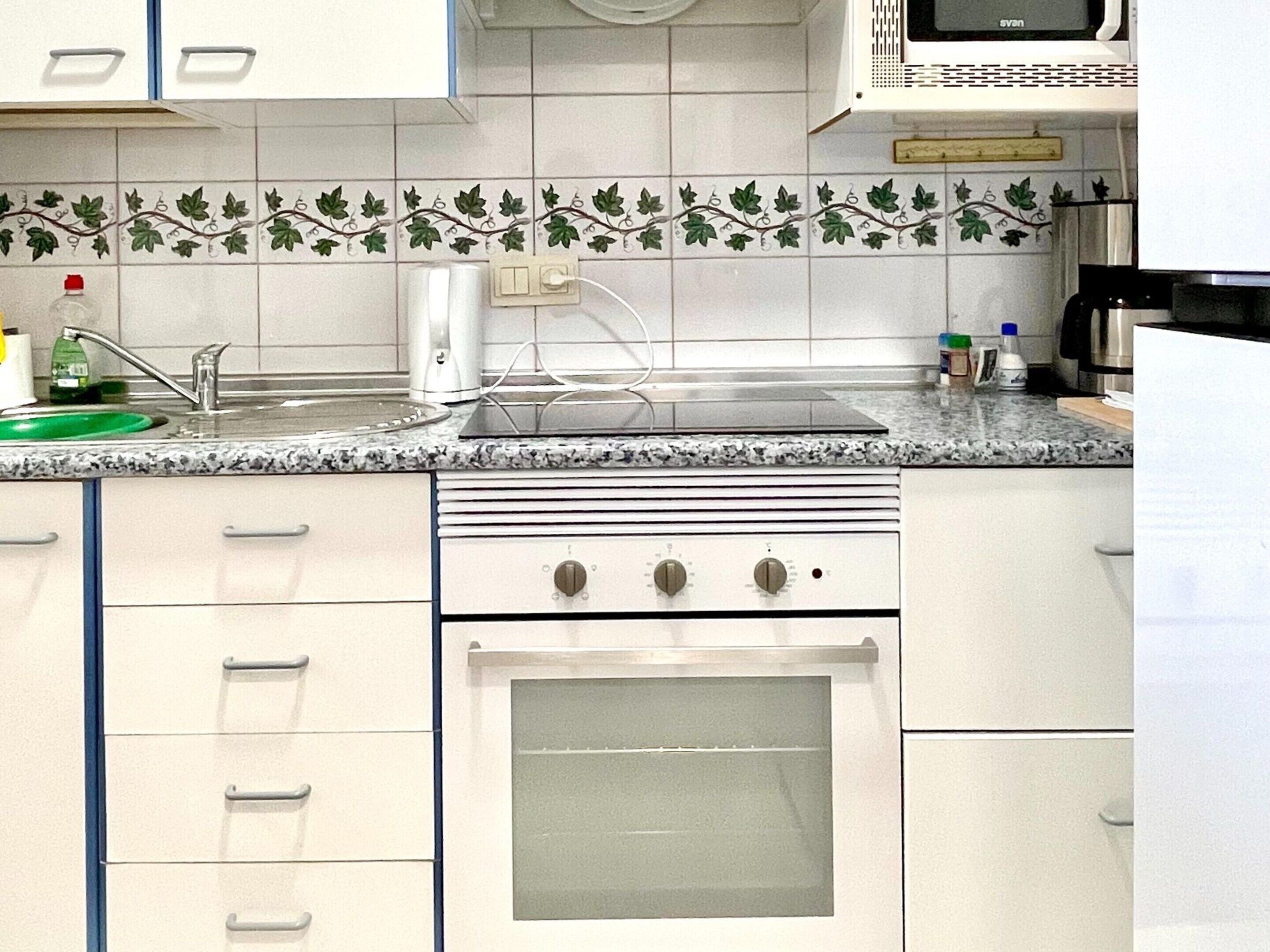 A kitchen with white cabinets, a stove, an oven, a microwave, and a tiled backsplash with a vine pattern.