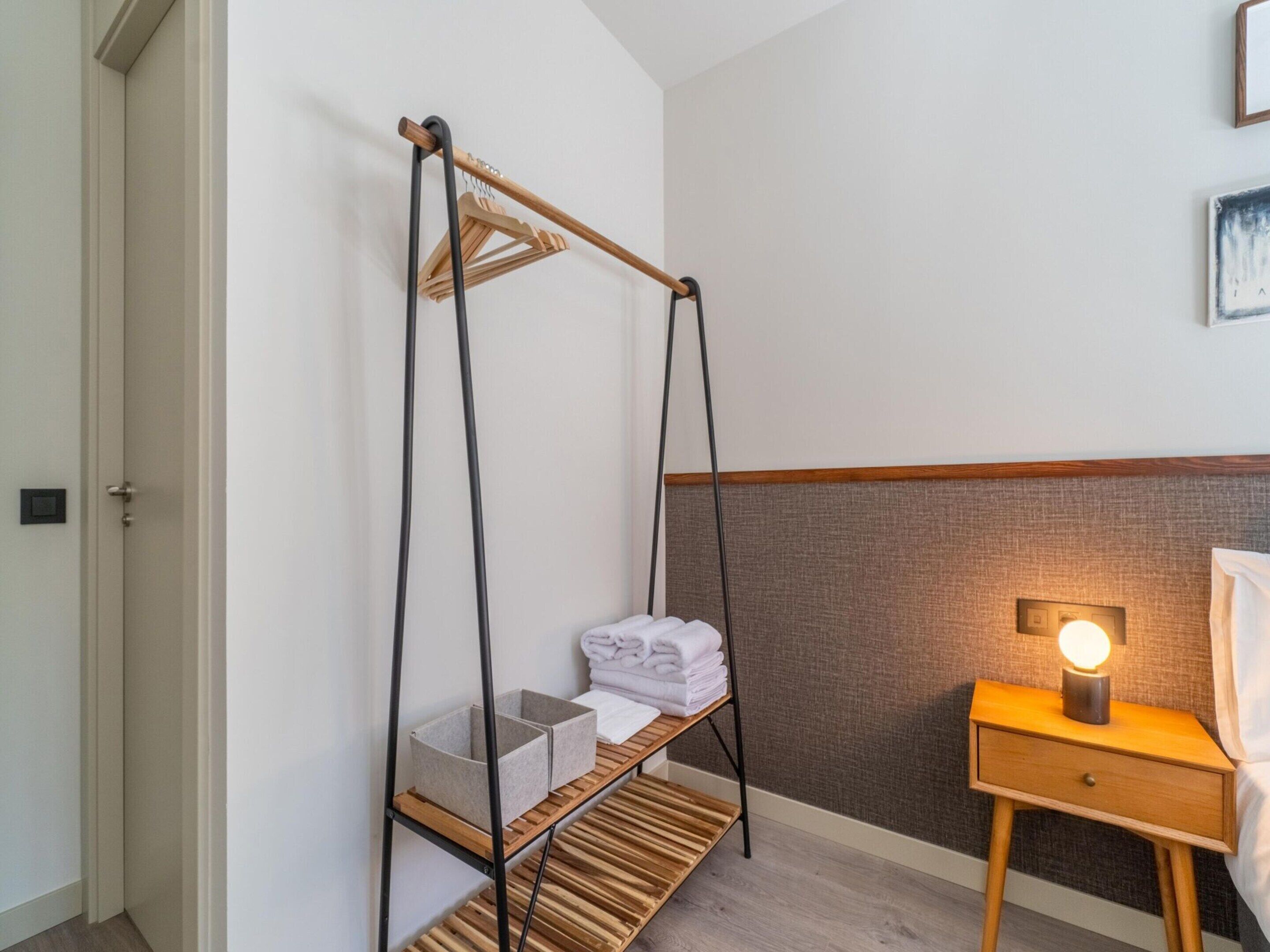 A hotel room with a wooden bed frame, a bedside table with a lamp, a hanging laundry rack, and a small shelf with folded towels.