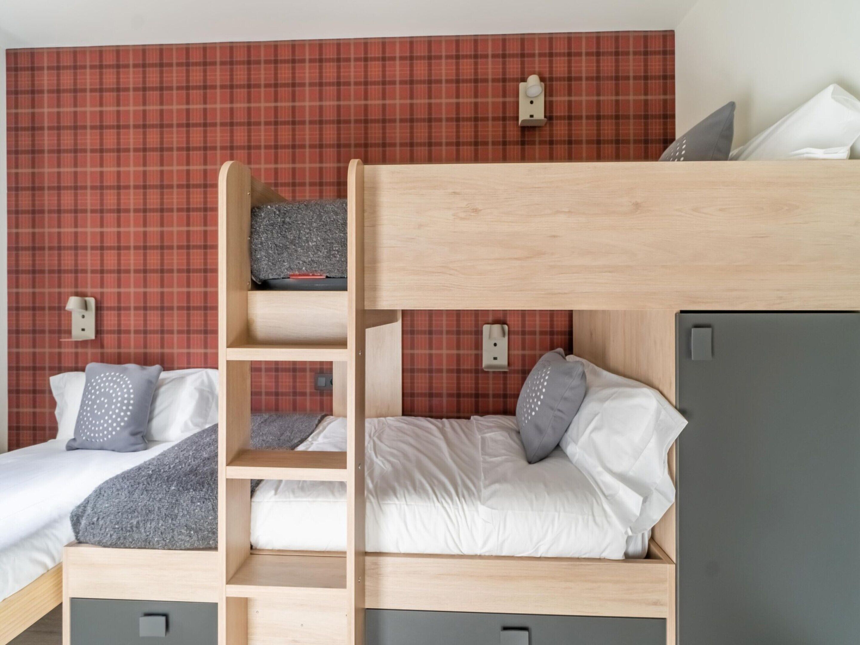 A bunk bed room with a red plaid wallpaper, two bedside tables, and a grey wardrobe.