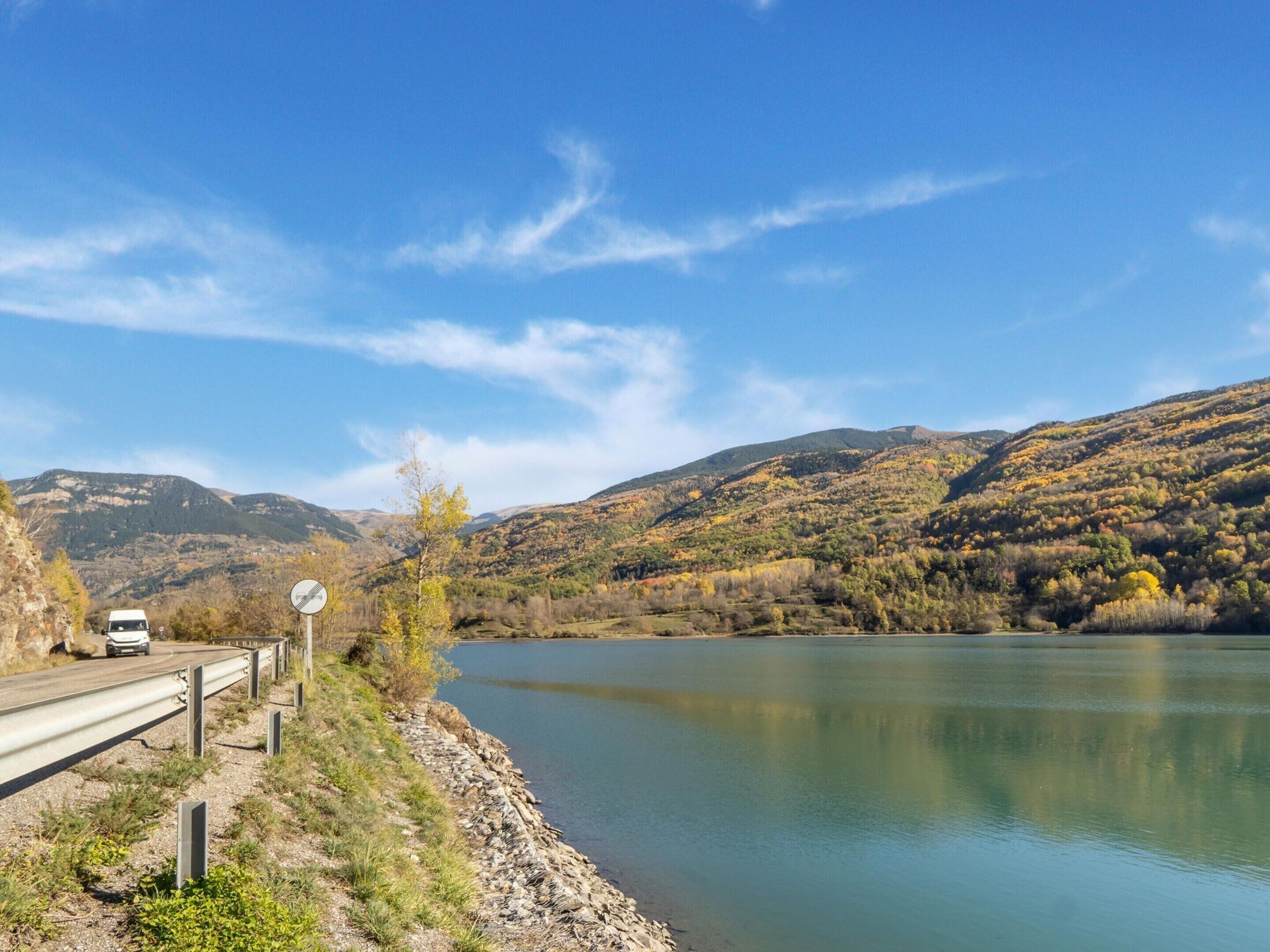 Una carretera con baranda de seguridad al lado de un lago tranquilo, rodeado de colinas con follaje otoñal.