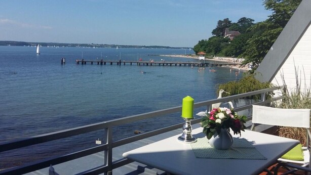 A table with a candle and flowers on a balcony overlooking a calm sea with boats and a pier.
