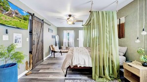 A bedroom with a bed, a desk, a chair, a ceiling fan, and a view of a house through a sliding door.