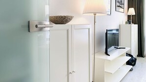 Moderni hotellihuone, jossa on lasiovi, valkoinen kaappi, jossa on punottu kori, lattiavalaisin, taulutelevisio ja pieni työpöytä, jonka päällä on laukku.