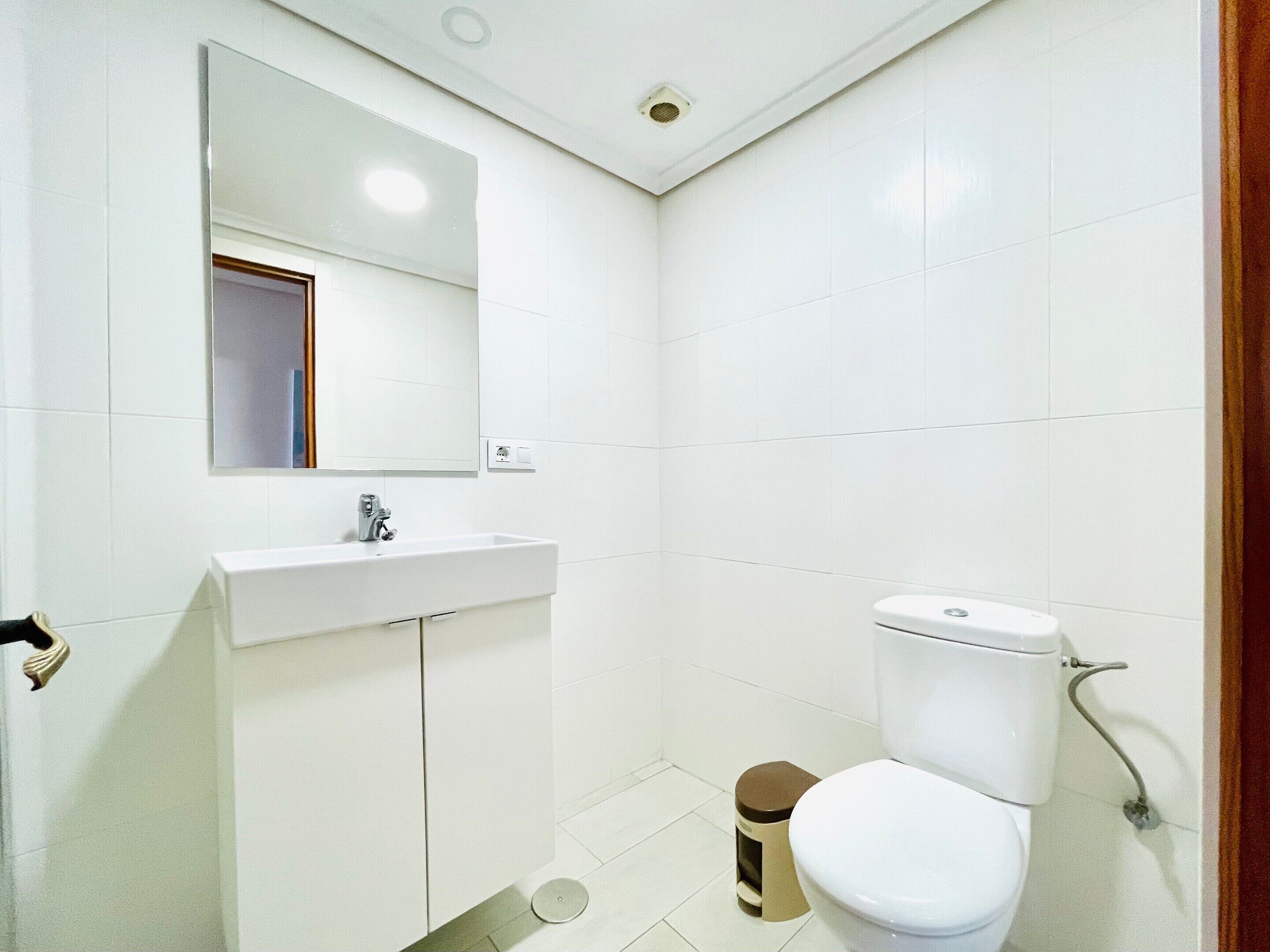 A small, clean bathroom with white tiles, a sink, a toilet, and a mirror.