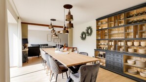 Een ruime eethoek met een lange houten tafel en moderne stoelen, een inbouwkeuken met houten kasten en hanglampen.