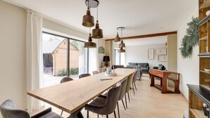 Een ruime eetruimte met een lange houten tafel en moderne stoelen, een gezellig zithoekje met een bank en een salontafel, en uitzicht op een houten gebouw buiten.