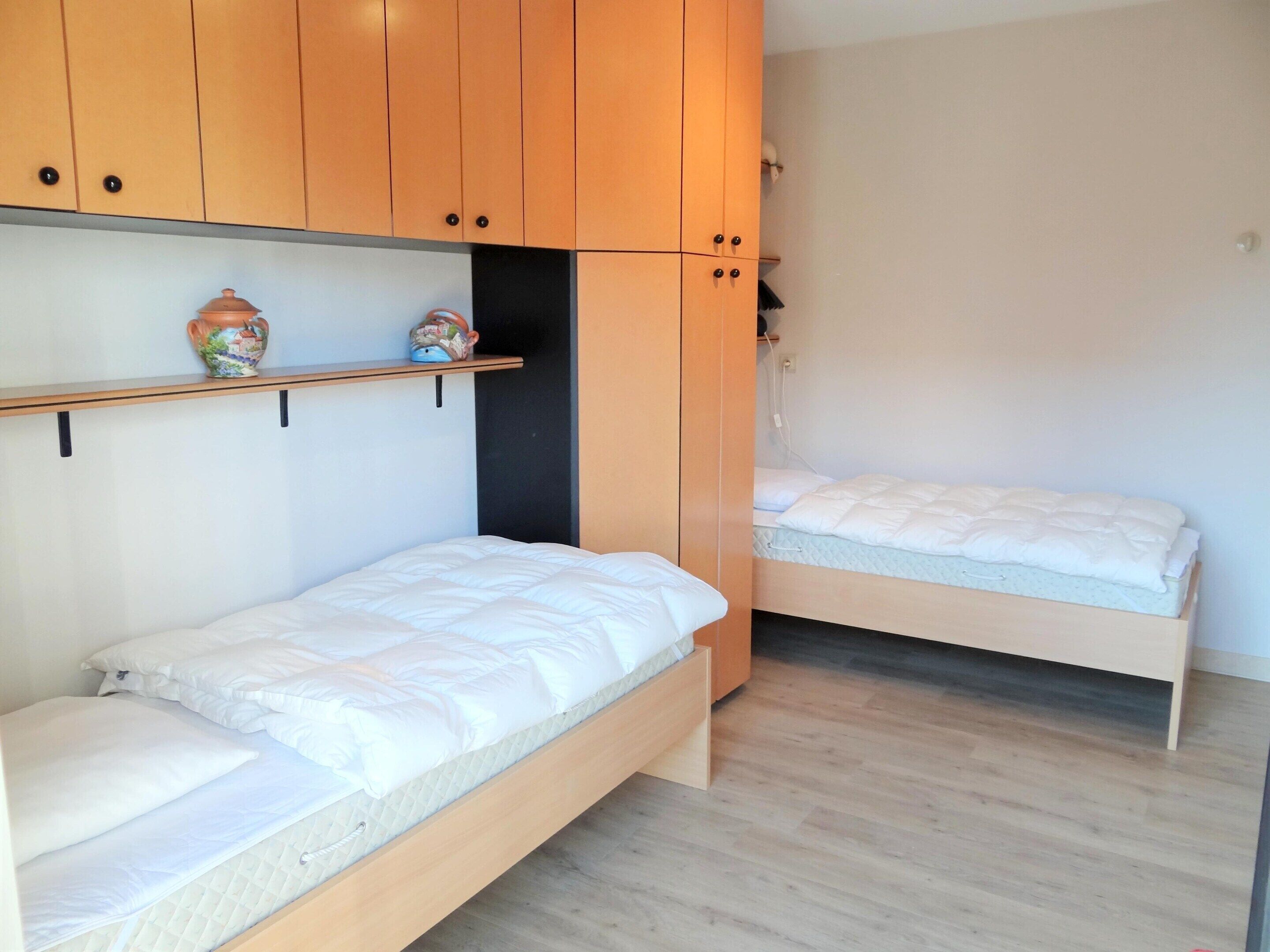 Quarto com duas camas de solteiro, armários de madeira e uma estante com objetos decorativos.