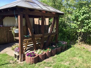 Ein hölzerner Pavillon mit geneigtem Dach, umgeben von Blumenbeeten mit verschiedenen Blumen.