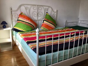 Ein Bett mit weißem Metallgestell, bunte, gestreifte Bettwäsche und zwei Kissen mit Streifen.