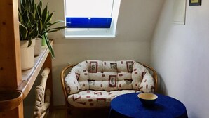 A cozy room with a high ceiling, a window with a view, a wicker chair, a small table with a bowl, and a potted plant. - Ferienwohnung in Klutz an der Ostsee (Klütz)