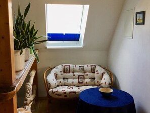 Ein gemütliches Zimmer mit hohen Decken, einem Fenster mit Aussicht, einem Korbstuhl, einem kleinen Tisch mit Schale und einer Topfpflanze.
