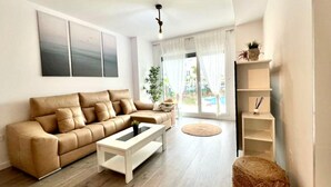 A modern living room with a beige sofa, a white coffee table, and a round rug. There is a large window with sheer curtains, a plant on a shelf, and a ceiling light. - Belvilla by OYO Playa Granada Beach & Golf 9 (Motril)