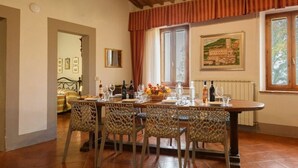 A dining area with a wooden table set for four, surrounded by patterned chairs. A window with curtains is visible, and a painting hangs on the wall. - Villa Colombini Comfortable Holiday Residence (Trequanda)