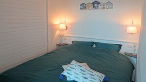Ein ordentlich bezogenes Bett mit einer grünen Tagesdecke und einem Kissen mit Fischmuster. Zwei Nachttischlampen und eine Wandleuchte sind vorhanden.