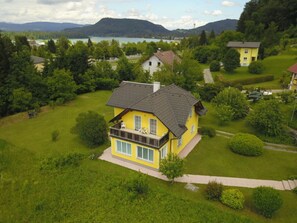 A yellow house with a balcony, surrounded by lush greenery and a lake in the background. - Holiday Home Anna Comfortable Vacation Home (Finkenstein am Faaker See)