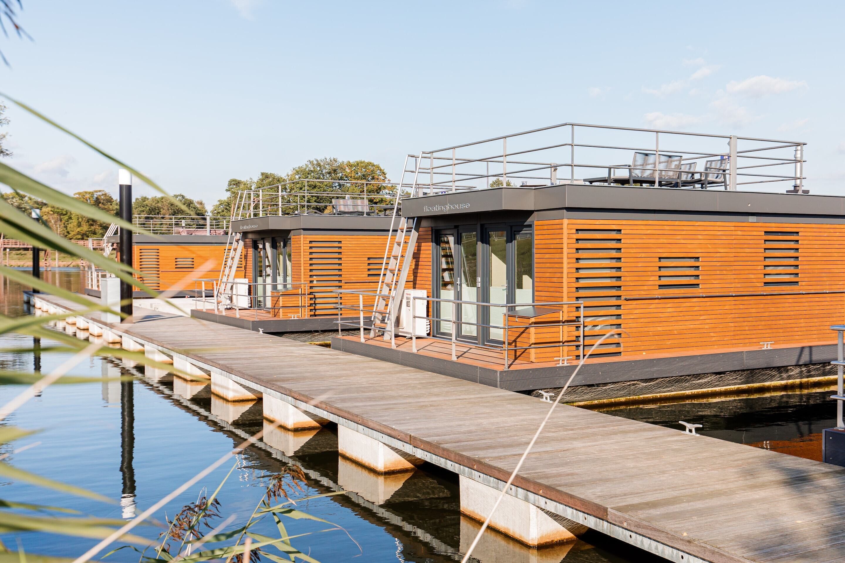 Un hotel flotante con fachada de madera, un espacio exterior cubierto y un muelle que se adentra en el agua.