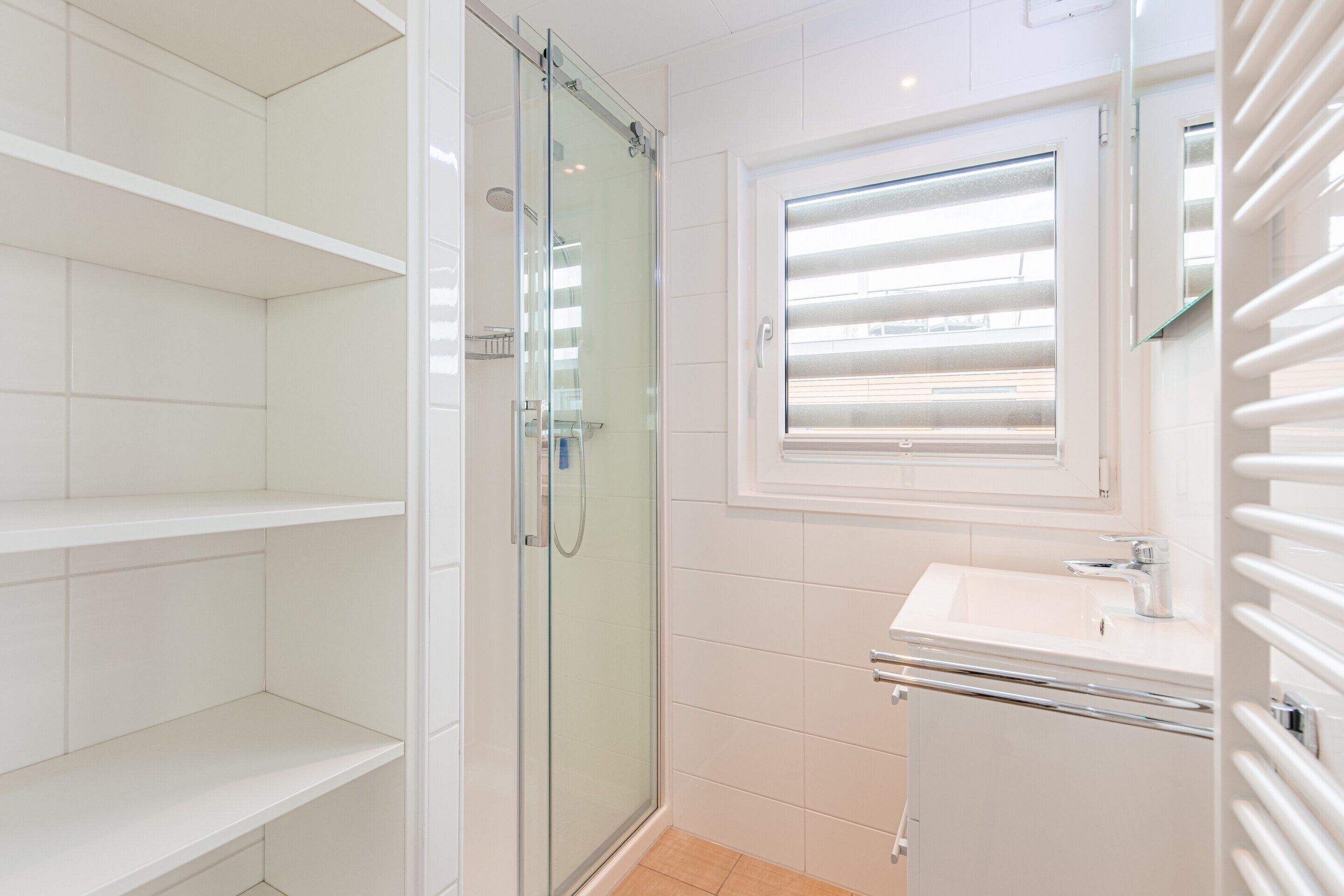 Une salle de bain moderne avec une douche à parois de verre, un lavabo blanc et une fenêtre équipée de stores.