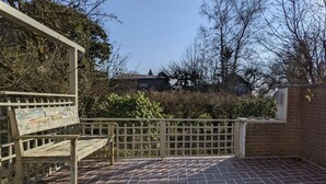 Une terrasse dotée d’un banc en bois et d’un sol en briques, entourée d’une clôture en bois et de plantes.