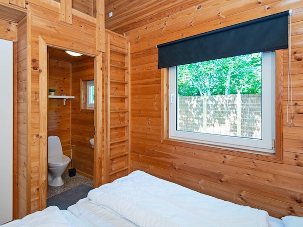 A wooden cabin room with a bed, a window with a view of trees, and a separate bathroom with a toilet and a small window.