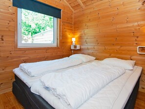 Un lit double avec une literie blanche, dans une chambre de chalet en bois dotée d’une fenêtre et d’une lampe fixée au mur.