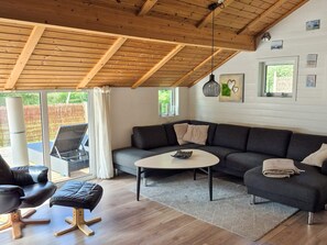 Un salon moderne avec un plafond en bois, un canapé gris, un fauteuil en cuir noir, une petite table basse et une vue sur l’extérieur grâce à de grandes fenêtres.