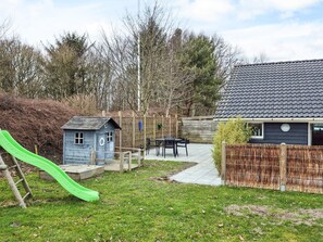Un jardin avec une petite maison de jeu, un toboggan, une terrasse avec des meubles d’extérieur et une maison dont le toit est de couleur sombre.