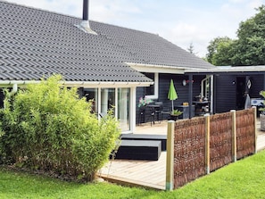 Ein Haus mit dunkler Fassade, einem Ziegeldach, einer Holzdielenveranda und einem umzäunten Gartenbereich.