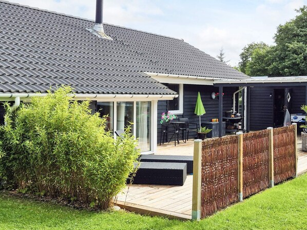 Une maison à l’extérieur sombre, avec un toit en tuiles, une terrasse en bois et un jardin clos.