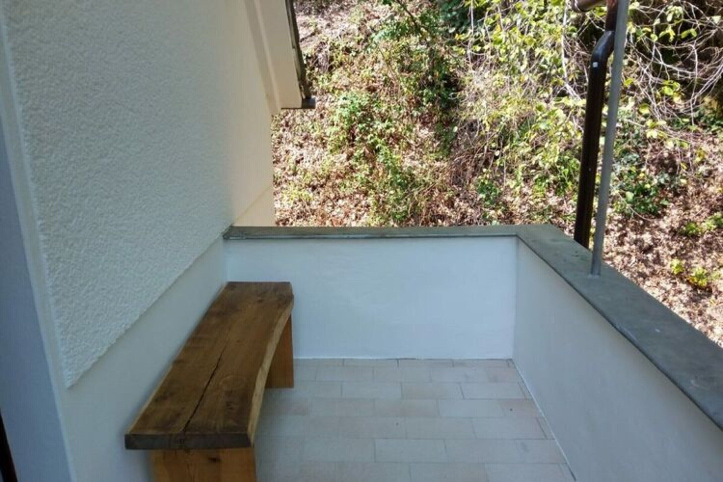 A small, enclosed balcony with a wooden bench, white walls, and a view of a garden with bushes and trees.