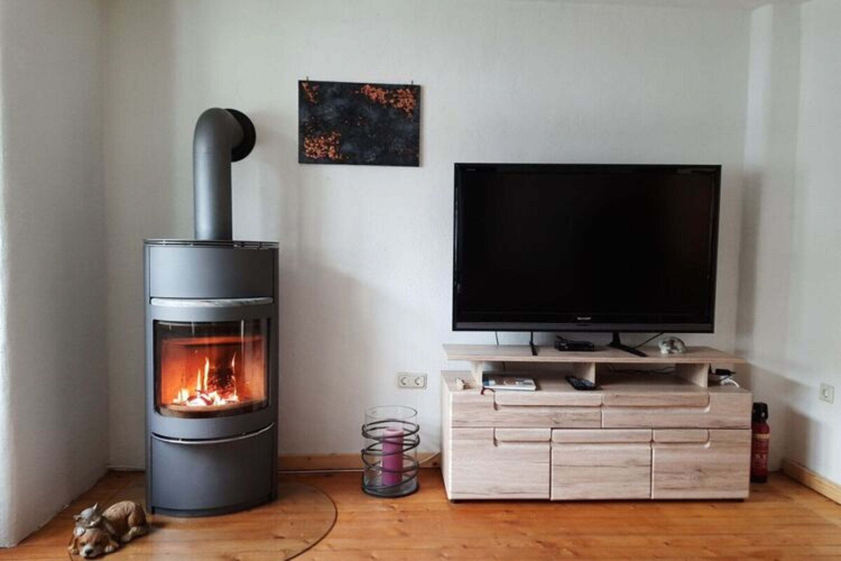 A modern living room with a wood-burning stove, a flat-screen TV on a wooden stand, and a small table with a candle holder.