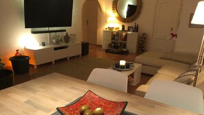 A modern living room with a wooden dining table, a flat-screen TV, a sofa, and a decorative mirror.