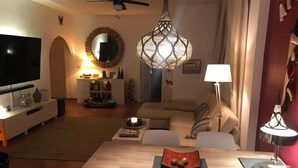 A modern living room with a flat-screen TV, a dining table with chairs, a sofa, decorative lamps, and a ceiling fan.