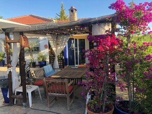 Une terrasse extérieure avec une table et des chaises en bois, entourée de plantes en pot et agrémentée d’une pergola illuminée de guirlandes lumineuses.