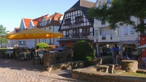 Uma praça de pedra com mesas ao ar livre, edifícios tradicionais em estilo de enxaimel e uma placa de hotel visível.