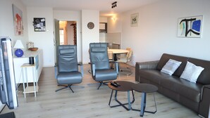 Una sala de estar moderna con un sofá de cuero marrón, dos sillones grises, una mesita redonda y un televisor sobre un mueble bajo.