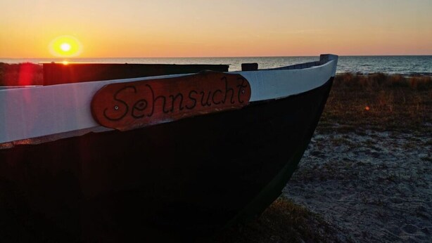 Ein Boot mit einem Holzschild, auf dem 'Sehnsucht' steht, am Strand bei Sonnenuntergang.