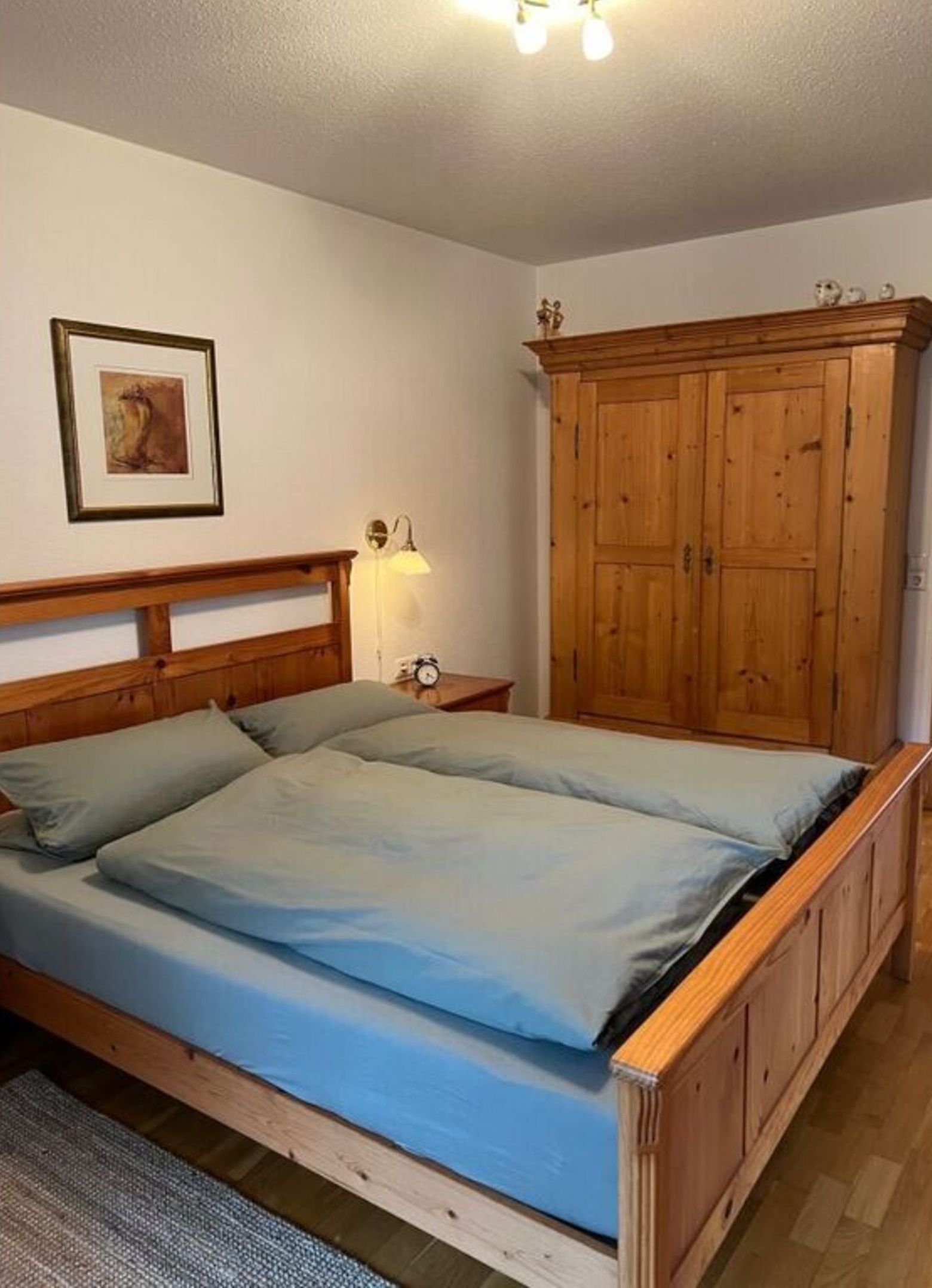 A bedroom with a wooden bed frame, a nightstand with a lamp, a wardrobe, and a framed picture on the wall.