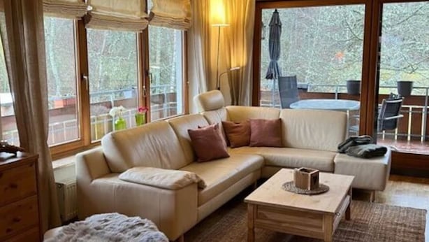 Una sala de estar acogedora con sofá, reposapiés y una mesa de centro, ventanas amplias con cortinas y vistas al exterior.